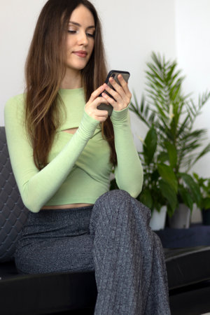 A beautiful, slim young woman sits with her phone in her hands and stares at the screen in front of houseplantsの写真素材