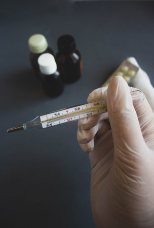 Hand holding a mercury thermometer and medicine bottles on a dark background