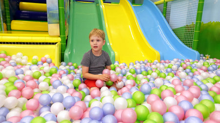 Blonde little boy 4-6 years old is sitting on multi coloured plastic balls in big dry paddling pool...