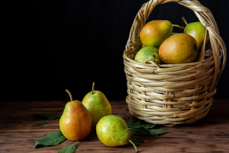 A wicker basket with ripe pears stands on a wooden table. Nearby are three pears.の写真素材