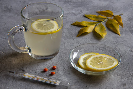 Ginger tea with a slice of lemon, lemon on a plate, a thermometer and pills, yellow leaves on a gray...