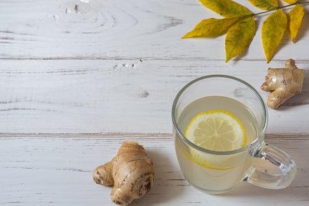 Ginger tea with a slice of lemon, yellow leaves of ginger root are on a white table. There is a copy...