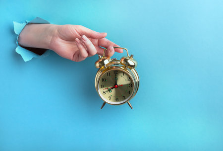 Female hand holds a golden alarm clock through a hole on a blue background. Time concept with place for text.の写真素材
