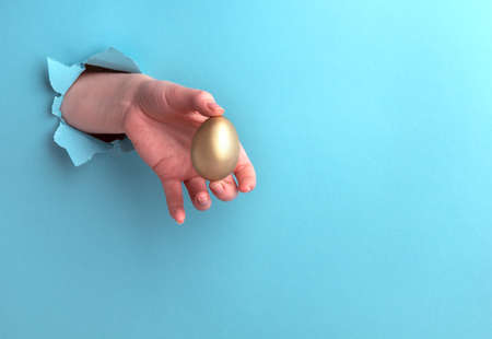 Golden egg in a female hand on a blue background with a hole. Horizontal orientation with copy space.の写真素材