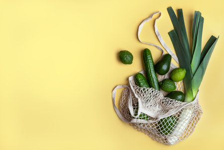 Green products in a reusable mesh bag on a yellow background. Environmental concept, top view, copy...