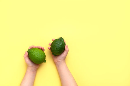 Children's hands hold two limes on a yellow background. Top view, copy space.