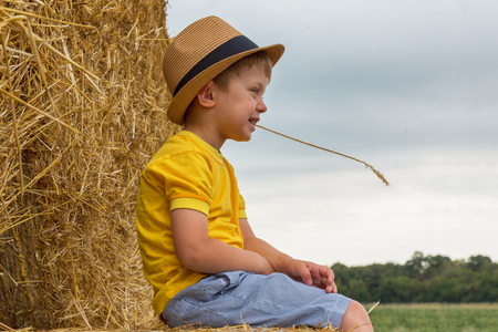 Little nice boy dressed in a yellow shirt and denim shorts and modern hat sits on a haystack in a summer day. Harvesting.の写真素材