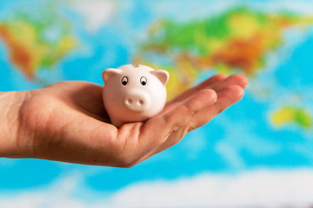 A tiny piggy bank is held in the hand. A colorful map of the world in the background. Travel...