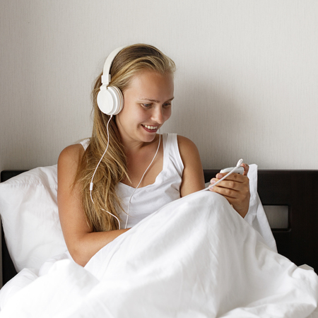 Young blond and happy smiling woman listen music and drink coffee in white bed in morning time.の写真素材