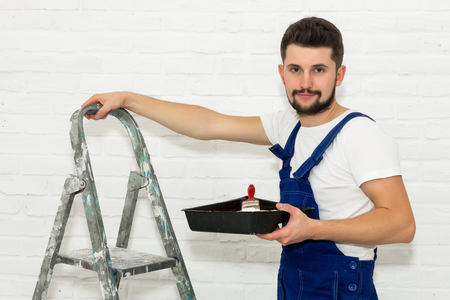 Young man with dark hair is painting the wall in white in his apartment. In his hand he holds a paint pan and paint brush, leaning against the ladder. He looks at the viewer and smiles.の写真素材
