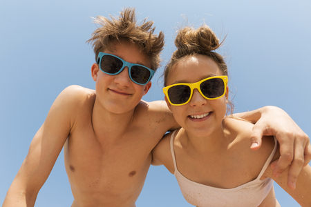 Two young and beautiful teenagers in colorful sunglasses hug on the beach against the blue sky.They are leaning towards the camera, looking at the viewer and laughing.の写真素材