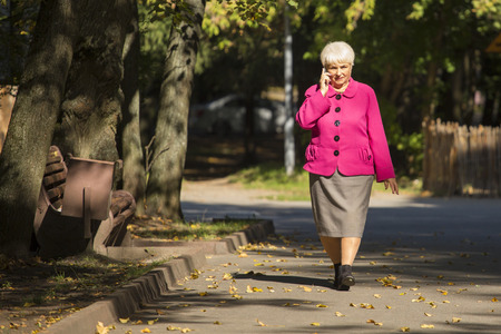A beautiful senior woman is walking and talking on the phone in a city park on sunny day.の写真素材