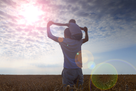 Happy family. Father and son on the wheat field. Man holding boy on shoulder. Looks at the sun. Back view.の写真素材