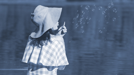 Young woman in a dress and a straw hat blowing bubbles against the background of the river. Back view, blue toned.の写真素材