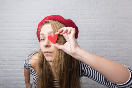 Funny young girl holding a red paper heart and laughing at the camera. Valentine's Day.