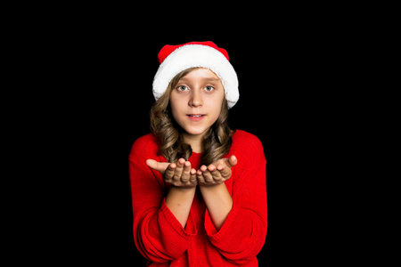 A young girl in a red sweater and a Santa Claus hat blows on her hands and looks at the viewer. Black isolate. Copy space.の写真素材