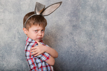 A funny, grumpy little boy dressed in a red checkered shirt and an old-fashioned hat, has a rabbit's...