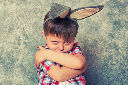 A funny, grumpy little boy dressed in a red checkered shirt and an old-fashioned hat, has a rabbit's ears on his head. Copy space.の写真素材