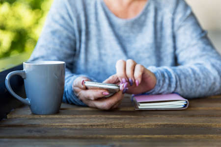 Elderly woman holding a cellphone, where to look for information. Next is a notebook and a cup of tea. Business concept. Close up.の写真素材