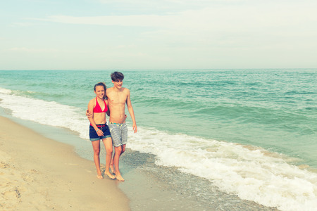 Two teenagers: a girl and a boy with blond hair, dressed in a swimsuit walks returning to the viewer on the sea beach.の写真素材