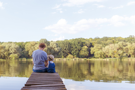 Dad and a tiny girl in dungarees relax at the river. Back view, copy space.の写真素材
