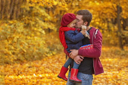 Father hugs and kisses his daughter in autumn day. The girl is holding a teddy bear. Copy space.の写真素材