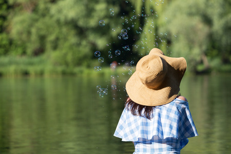 Young woman in a dress and a straw hat blowing bubbles against the background of the river. Back view.の写真素材