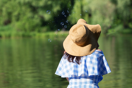Young woman in a dress and a straw hat blowing bubbles against the background of the river. Back view.の写真素材