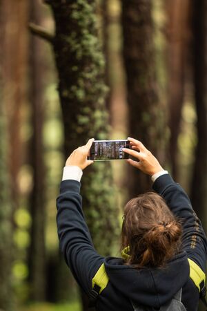 A young woman with brown hair taking picture of trees in the forest with her smartphone, medium shotの写真素材