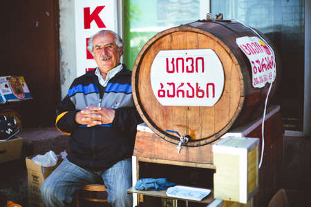 Tbilisi, Georgia - June 15, 2014: Elderly Georgian man selling a local drink in a barrel while sitting on a chair near his shop.のeditorial素材