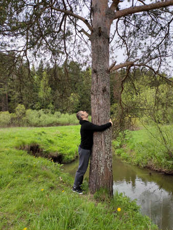 A young man is resting in the forest near a stream or lake. The man embraces a old tree on a forest glade. の写真素材
