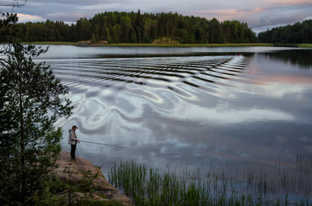 Fisher-woman standing on a rock with a fishing rod and fishing. On the lake waves diverge from the boat.の写真素材