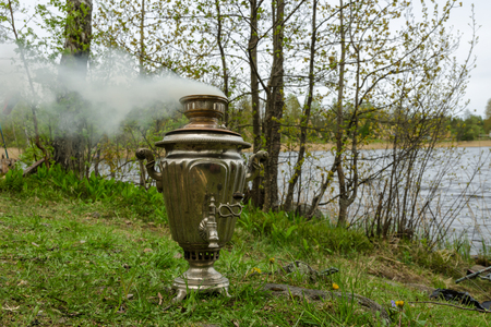 The samovar stands on the grass on the bank of the reservoir. The wind blows away the smoke from the samovar.の写真素材