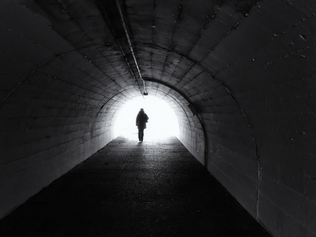 Woman goes from us to the light in the tunnel.  At the end of the tunnel you can see a bright light. A woman is dressed in winter clothes. Black and White.の写真素材