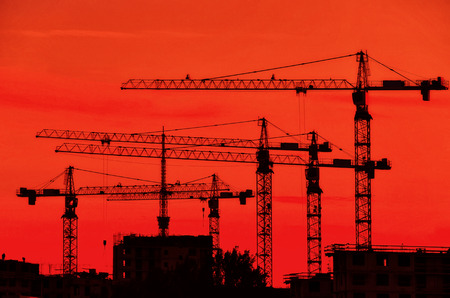Silhouettes of several construction cranes against the background of the evening red sky. There are buildings under construction underneath. Background.の写真素材