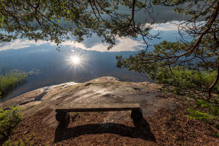 A simple wooden bench stands on the stone shore of the lake. There is a shadow from the bench and pine branches framing it. The water reflects the bright sun and the forest on the opposite bank.の写真素材