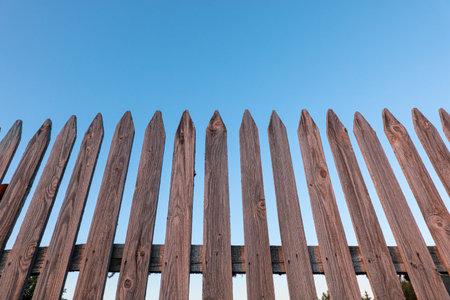 A look from below at an unpainted wooden fence made of pointed slats. The wooden elements are...