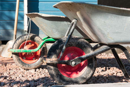 Two dirty wheelbarrows with silver bodies and red wheels sit on the gravel driveway. A building wall and a shovel are visible in the background. Close-up. Background.の写真素材