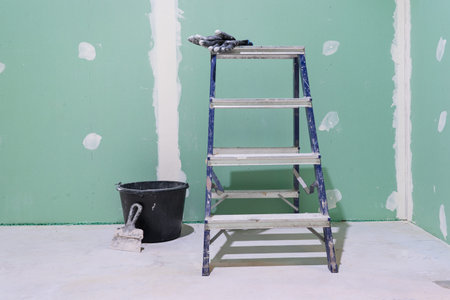 Fragment of a wall made of new green plasterboard. There is a construction ladder and a bucket of putty on the dirty floor. Gloves and spatula included. Background.の写真素材