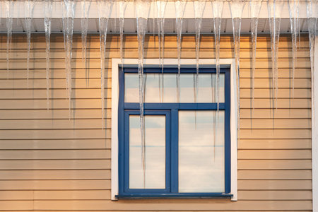 Fragment of a house wall made of beige siding with a window. Large icicles hang from the roof. There...