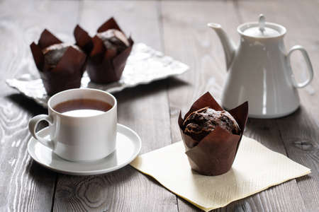 A cup of tea and dark cupcakes on a wooden table. The texture of the wood is visible, there is a white teapot. food. lifestyle.の写真素材