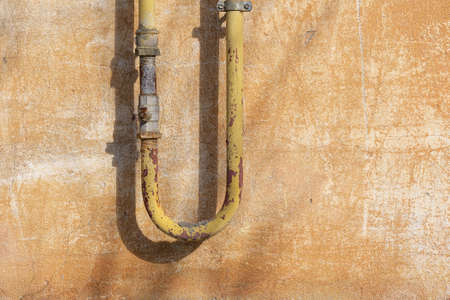 An old yellow U-shaped gas pipe against the background of a shabby plastered wall. There are stopcocks and rust on the metal. background. texture.の写真素材