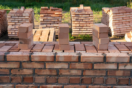 Frontal view of a traditional brick wall fence under construction. Pallets with red bricks are...