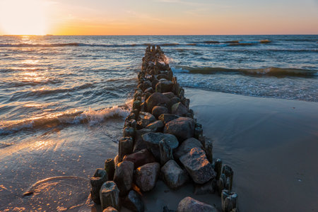 Perspective view of an old sea breakwater made of logs and stones. The waves coming from the sea are illuminated by the setting sun. Seascape.の写真素材