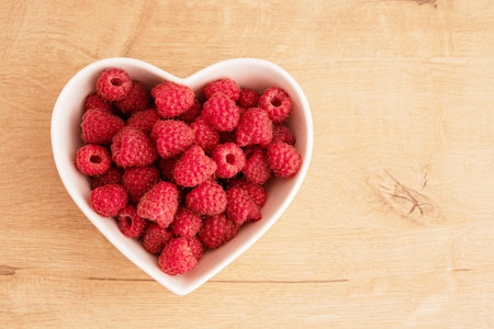 Fresh raspberries lie in a white heart-shaped salad bowl. There is a table surface made of light...
