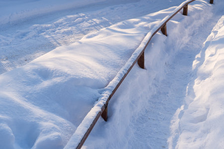 A fragment of a pedestrian path, trampled in deep snow and fenced with a metal barrier. A road is visible in the background.の写真素材