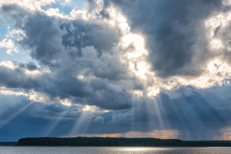The suns rays break through the dark clouds over the lake. A strip of forest is visible on the...