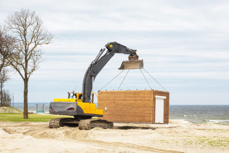 A yellow excavator transports a wooden pavilion along a sandy beach on the seashore in winter. Landscape.の写真素材
