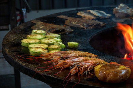 Large shrimp, potatoes, zucchini and flounder are cooked on the metal surface of the circular grill....