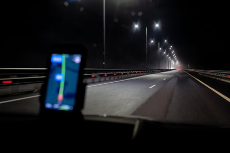 Dark night highway goes far ahead, red lights of cars are visible. There is a blurry smartphone with...
